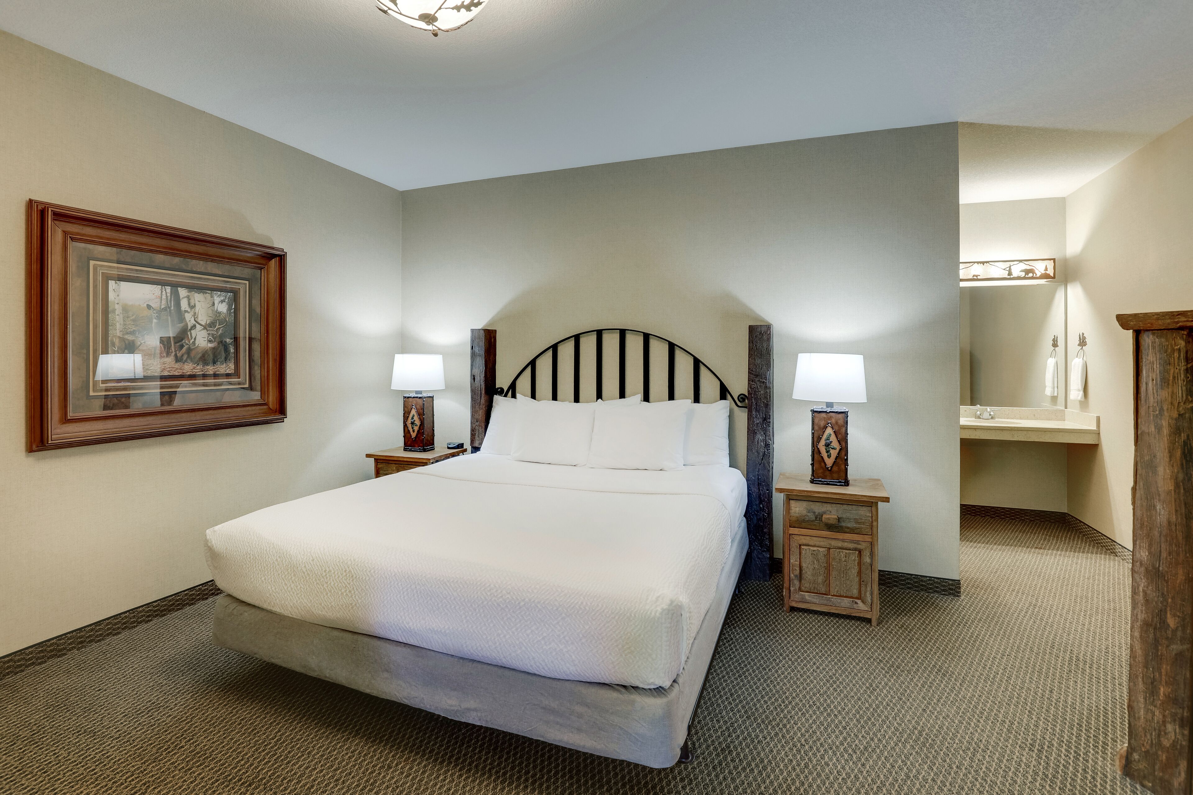 A neatly arranged hotel room with a large bed, two bedside tables with lamps, a framed picture on the wall, and a bathroom with a mirror and sink.