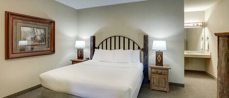 A neatly arranged hotel room with a large bed, two bedside tables with lamps, a framed picture on the wall, and a bathroom with a mirror and sink.