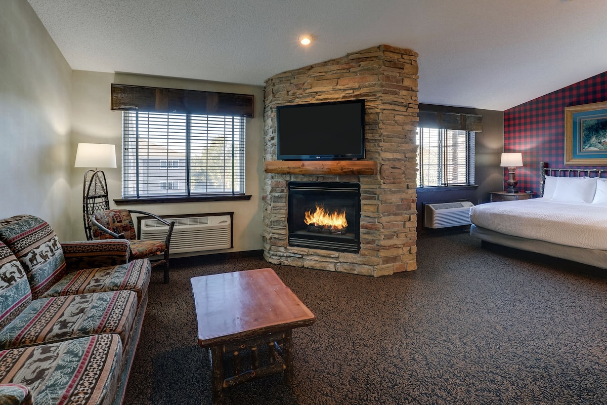 A cozy room with a stone fireplace, a flat-screen TV mounted above it, a sofa, a bed, and a window with blinds.