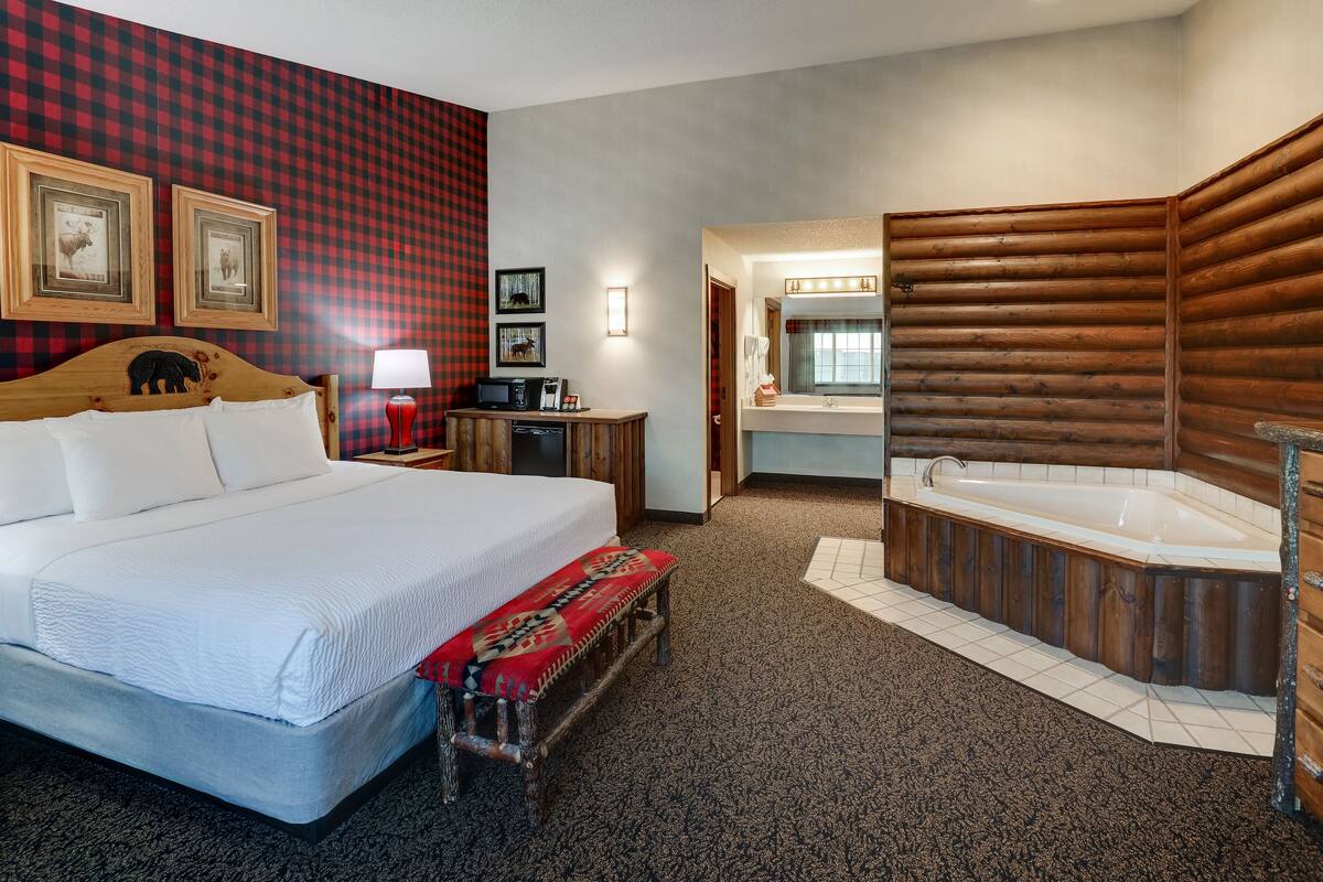 A hotel room with a large bed, a wooden bench, a desk with a lamp, a TV, and a bathroom with a wooden bathtub.