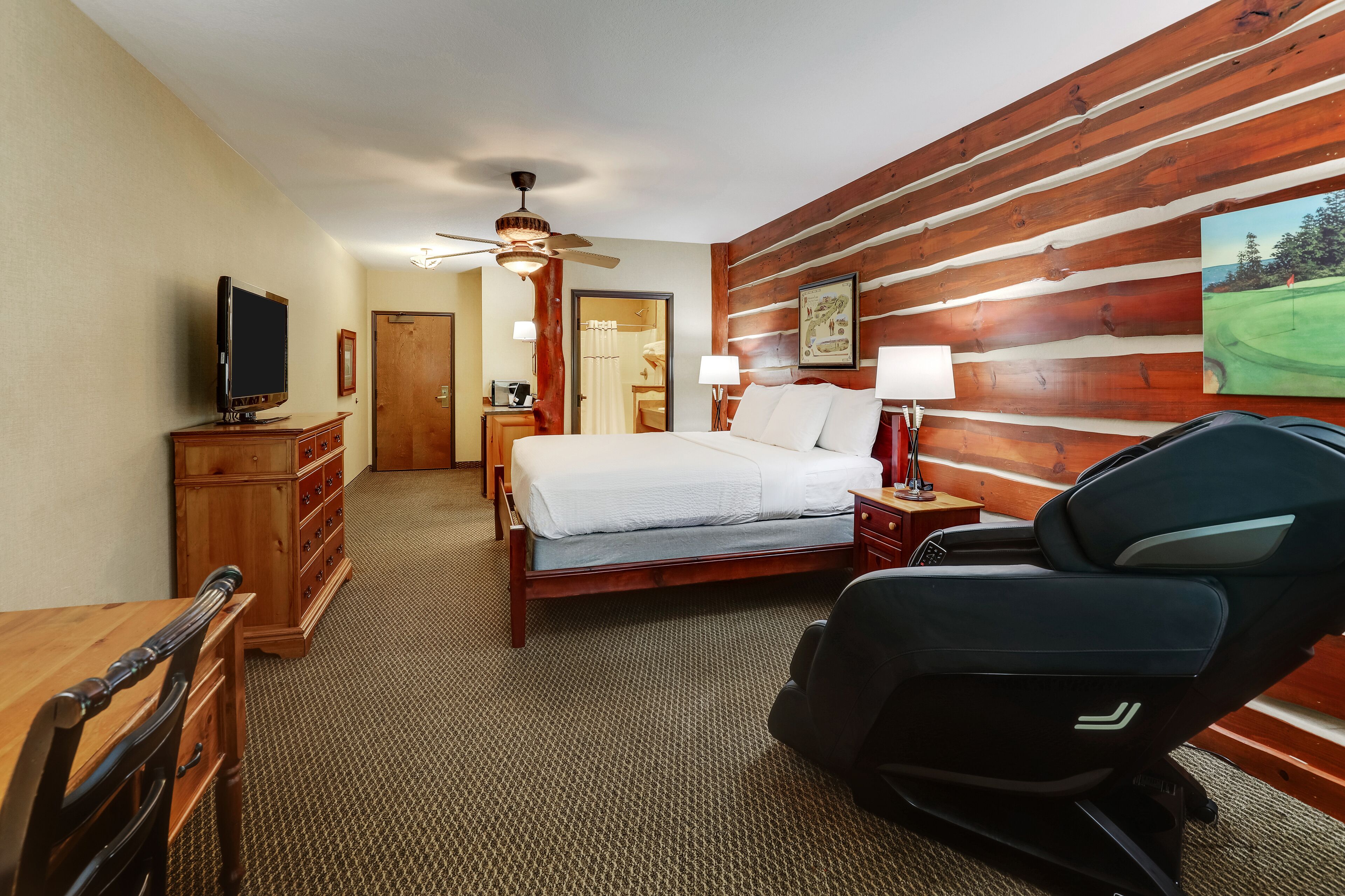 A hotel room with a bed, a desk, a chair, a television, a ceiling fan, and a wooden wall.