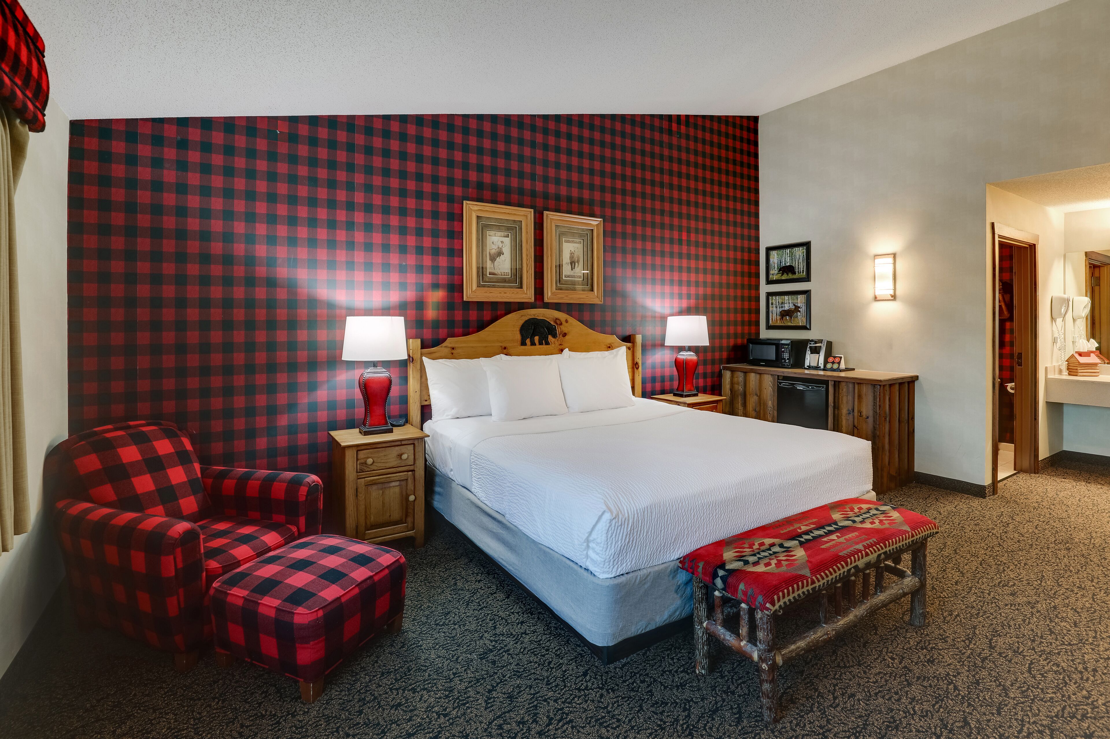 Une chambre d’hôtel avec un mur à carreaux, un lit doté d’une tête de lit en bois, une chaise à carreaux rouges et noirs, un petit banc en bois et une table de chevet avec une lampe.