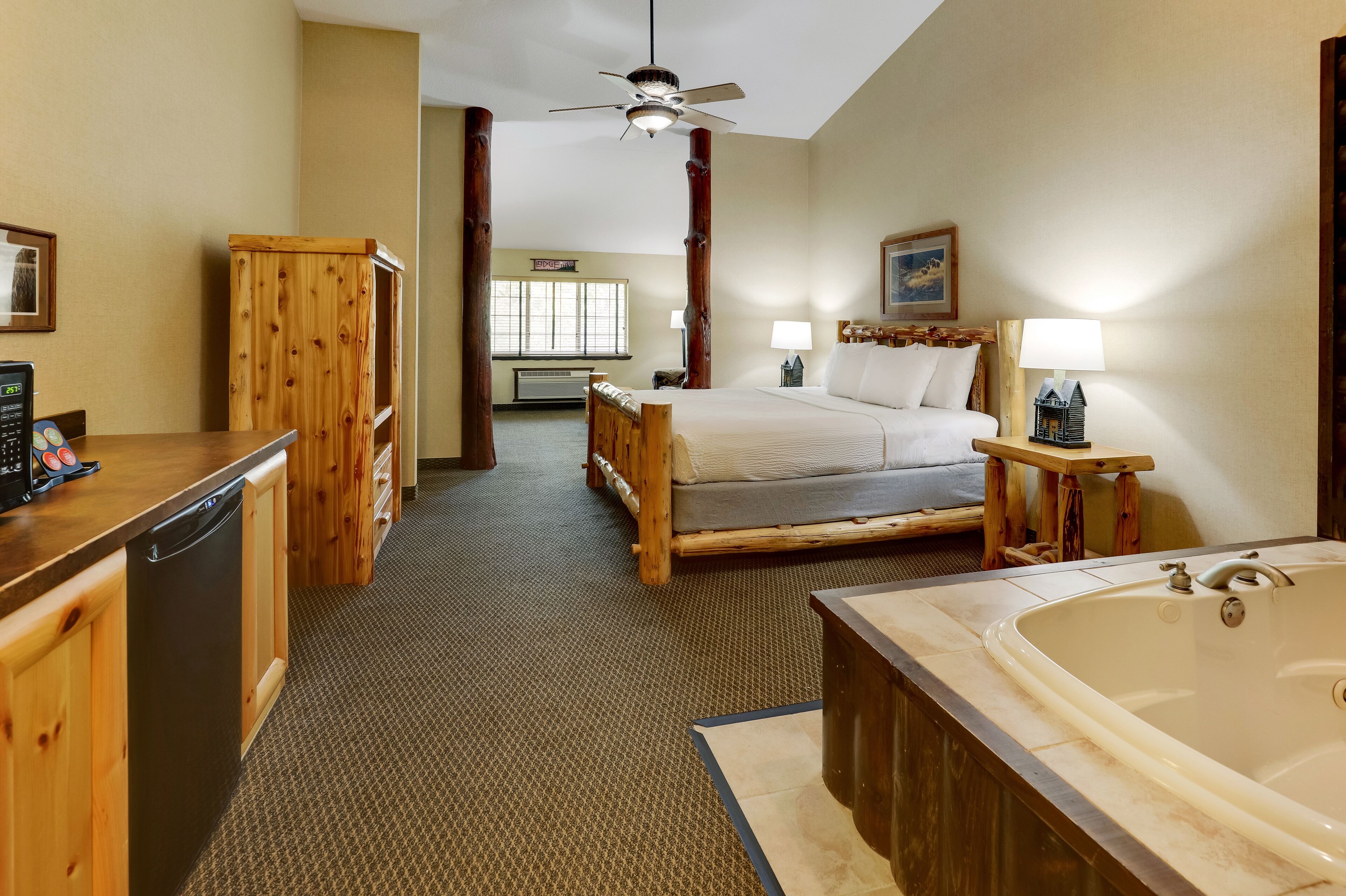 A hotel room with a wooden bed, a large bathtub, a wooden desk with a dishwasher, and a ceiling fan.