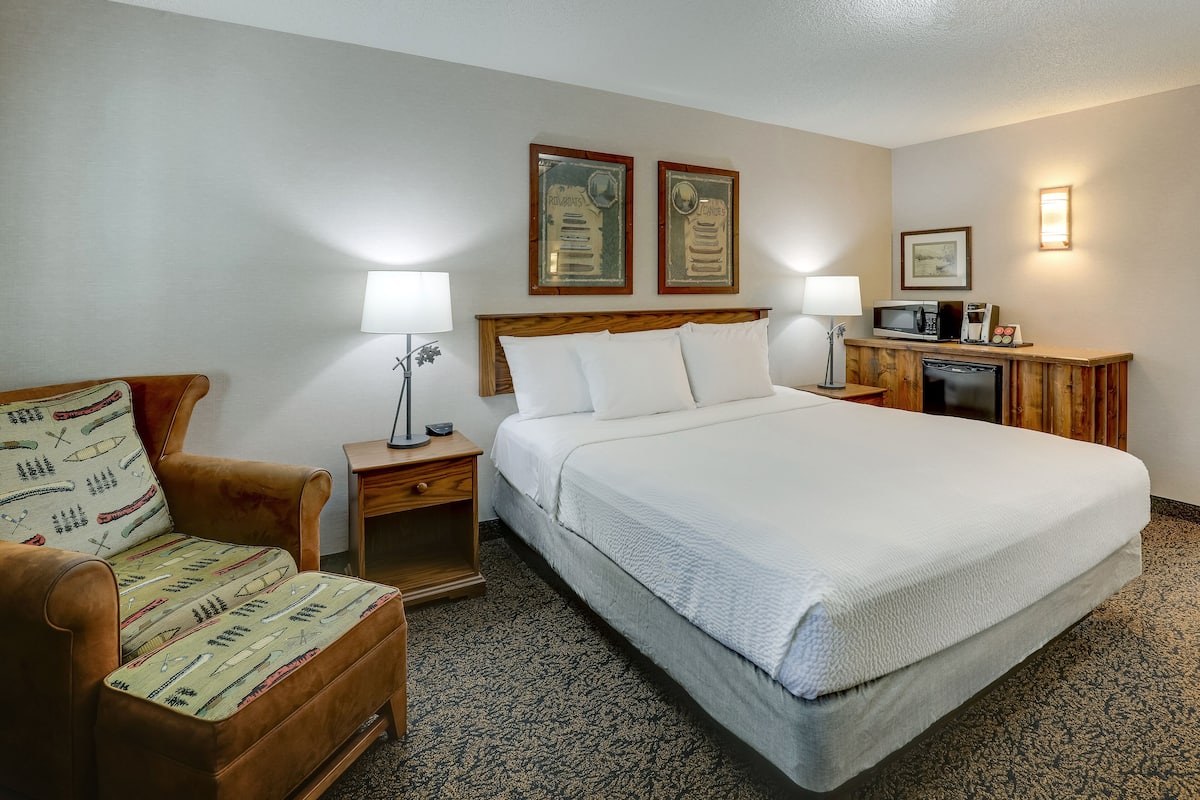 A hotel room with a bed, a wooden nightstand, a lamp, a chair with a patterned cushion, and a microwave on a wooden cabinet.