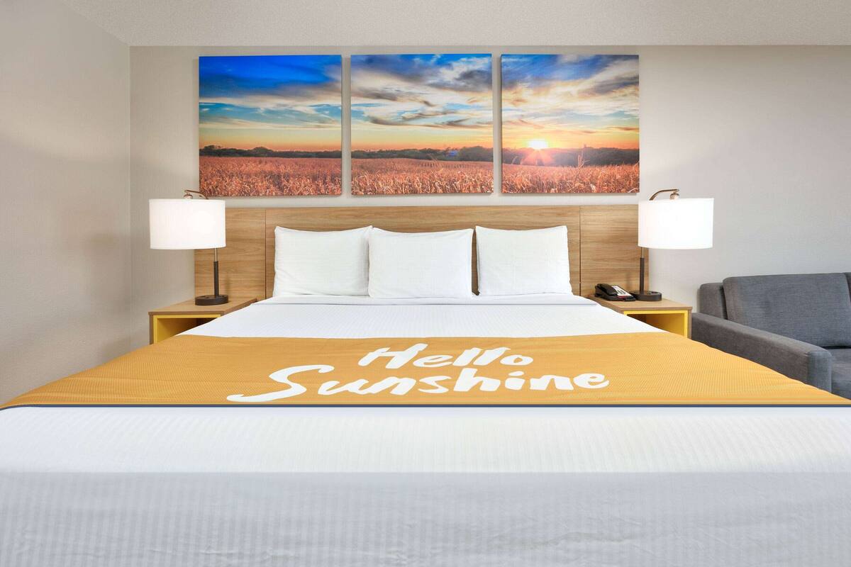 A hotel room with a bed, a headboard with a sunset view, two bedside lamps, a bedside phone, and a "Hello Sunshine" sign on the bedspread.
