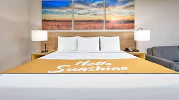 A hotel room with a bed, a headboard with a sunset view, two bedside lamps, a bedside phone, and a "Hello Sunshine" sign on the bedspread.