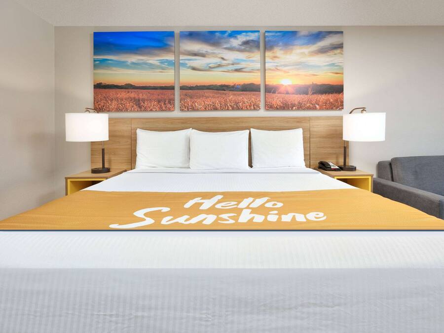 A hotel room with a bed, a headboard with a sunset view, two bedside lamps, a bedside phone, and a "Hello Sunshine" sign on the bedspread.