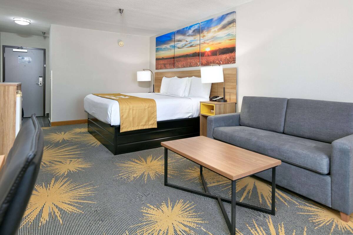 A hotel room with a bed, a gray sofa, a small table, and a painting of a sunset on the wall.