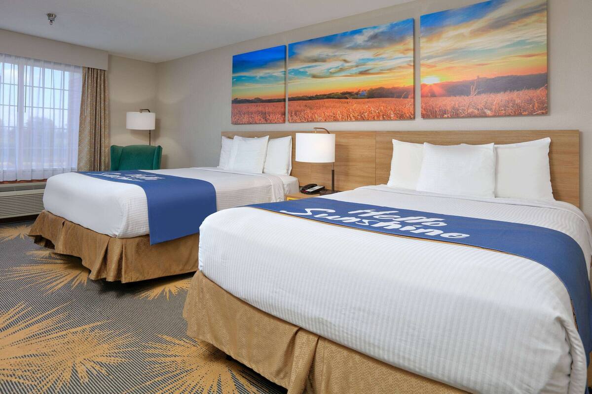 A hotel room with two beds, a large window with curtains, a wall-mounted lamp, and two landscape paintings.