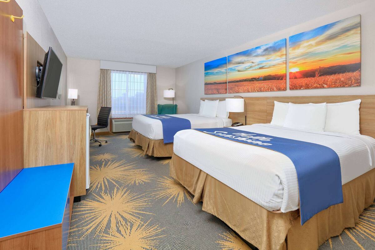 A hotel room with two beds, a desk, a TV, and a large window with a sunset view.