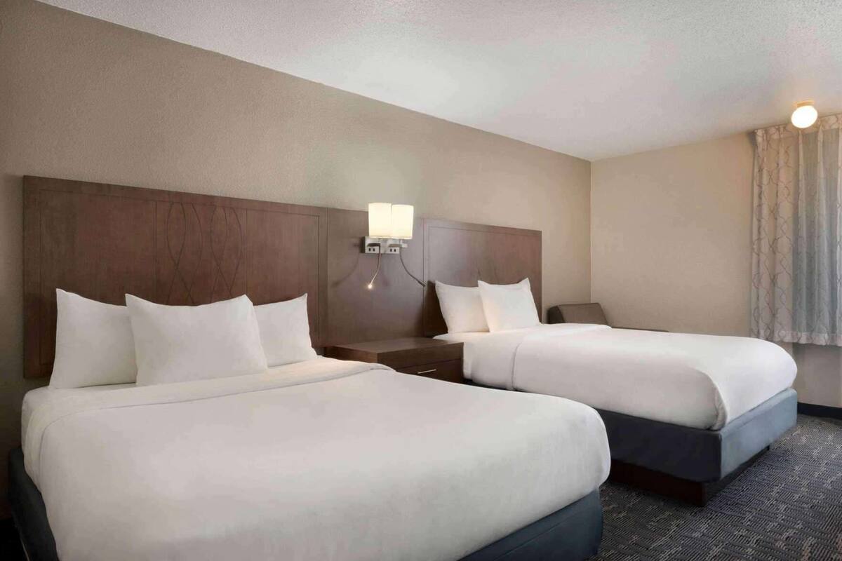 Une chambre d’hôtel avec deux lits, une table de chevet, une lampe fixée au mur et une fenêtre avec des rideaux.
