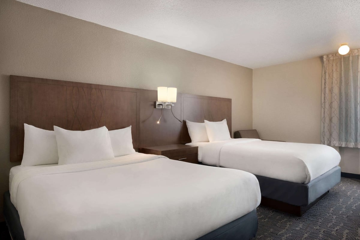 Une chambre d’hôtel avec deux lits, une table de chevet, une lampe fixée au mur et une fenêtre avec des rideaux.