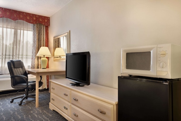 A hotel room with a desk, chair, television, microwave, and refrigerator.