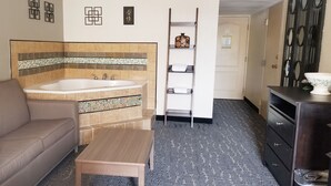 A hotel room with a beige sofa, a wooden table, a large bathtub, a shelf with decorative items, and a dark wooden dresser. - Baymont by Wyndham Des Moines North (Des Moines)