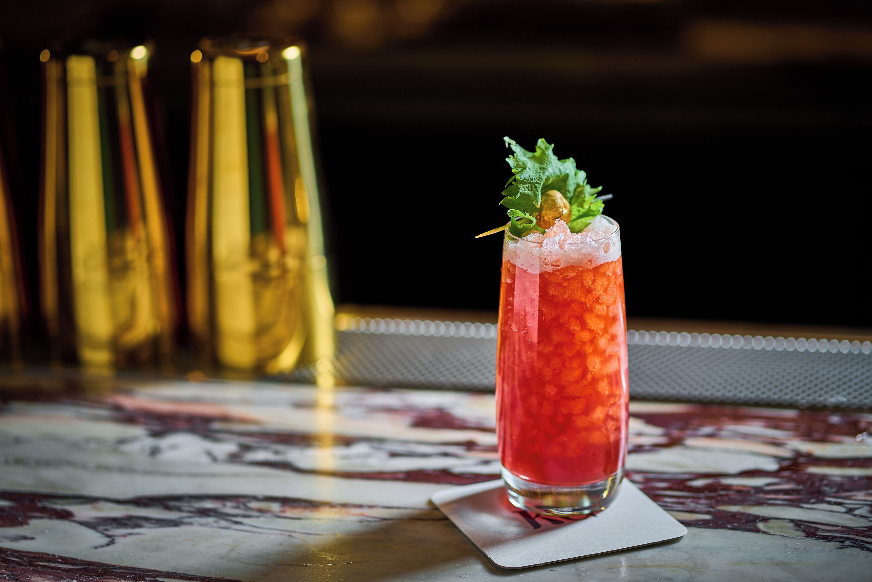 A tall glass with a red drink, garnished with a leaf and a nut, on a marble surface.