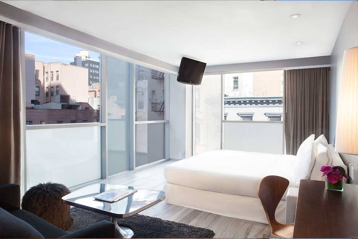 Quarto de hotel moderno com uma cama grande, uma mesa pequena, uma cadeira e uma TV fixada na parede.