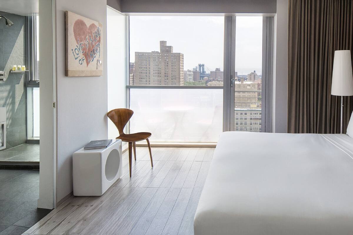 Quarto de hotel moderno com uma cama grande, uma cadeira, vista da cidade e uma janela com cortinas.