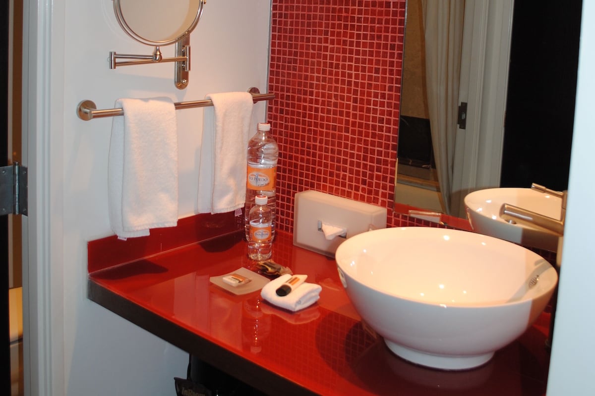 A bathroom with a red tiled wall, a white sink, a round mirror, and a towel rack.