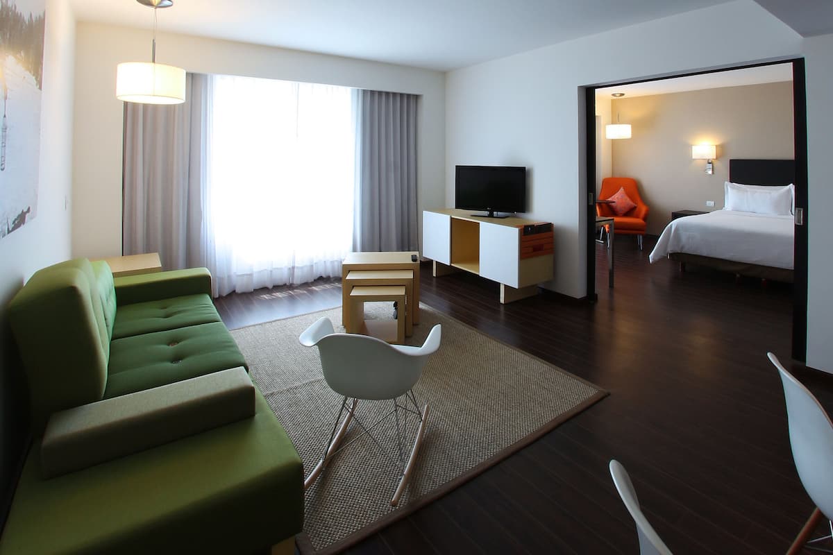 Habitación de hotel con sofá verde, una silla blanca, una pequeña mesa de madera, un televisor sobre un mueble y una cama con ropa blanca.