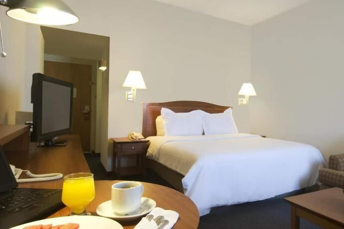 A hotel room with a bed, bedside table, lamp, and a desk with a computer and a glass of orange juice.