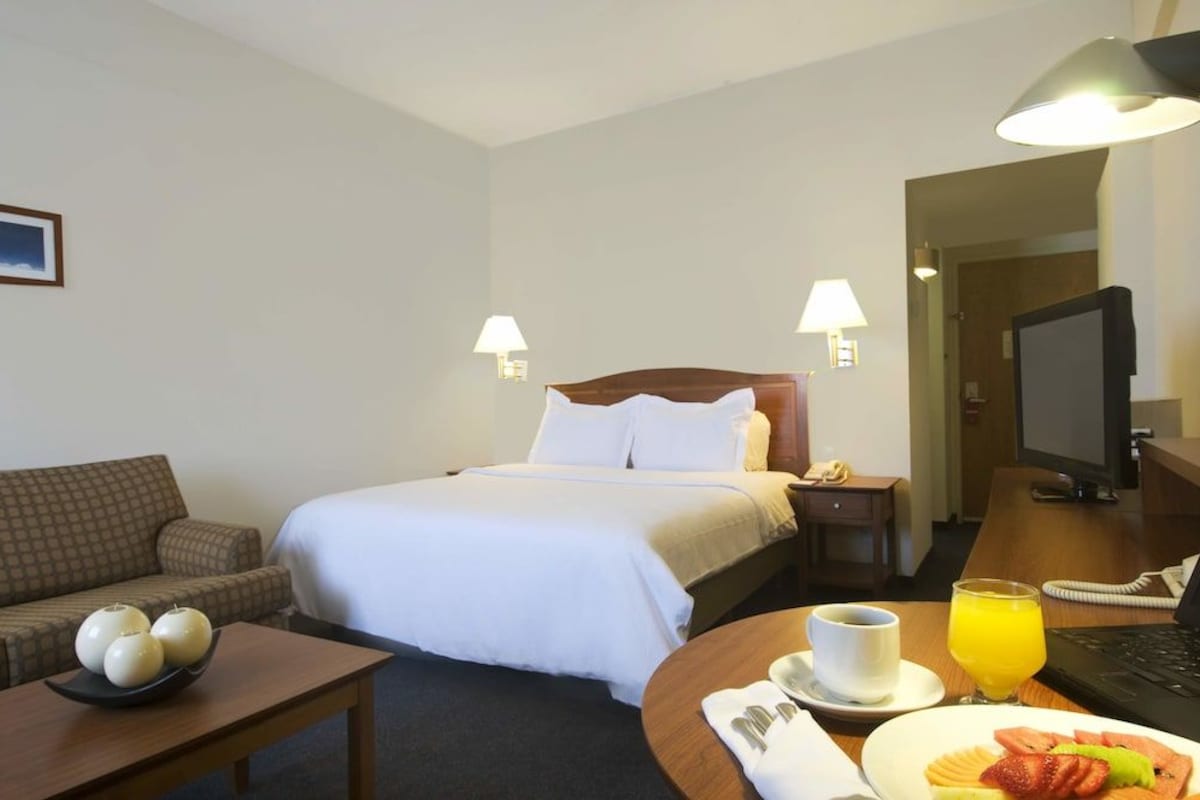 Una habitación de hotel con una cama, un escritorio con computadora, una silla, una mesita con un frutero y una lámpara en la pared.