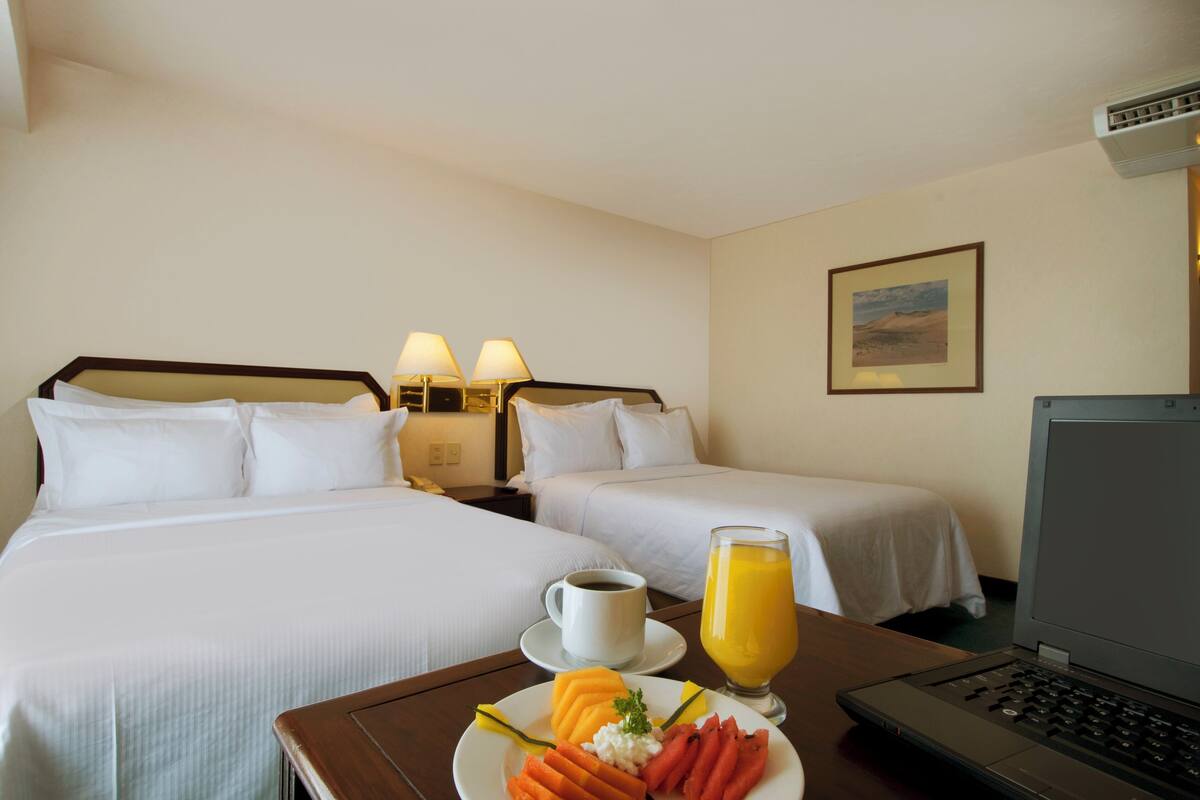 A hotel room with two beds, a laptop, a breakfast tray with fruit and coffee, and a painting on the wall.