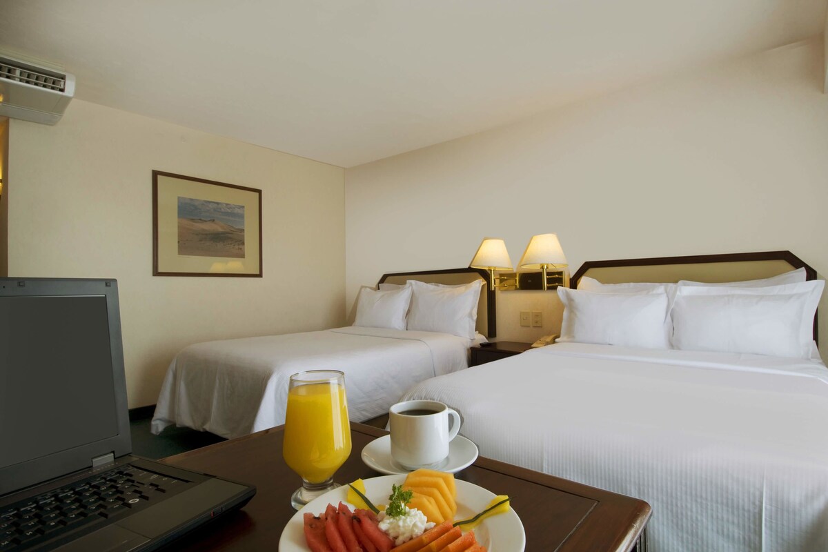 Habitación de hotel con dos camas, un escritorio con una computadora portátil, un plato de fruta, una taza de café y un vaso de jugo de naranja.