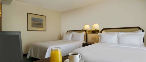 A hotel room with two beds, a desk with a laptop, a plate of fruit, a cup of coffee, and a glass of orange juice.
