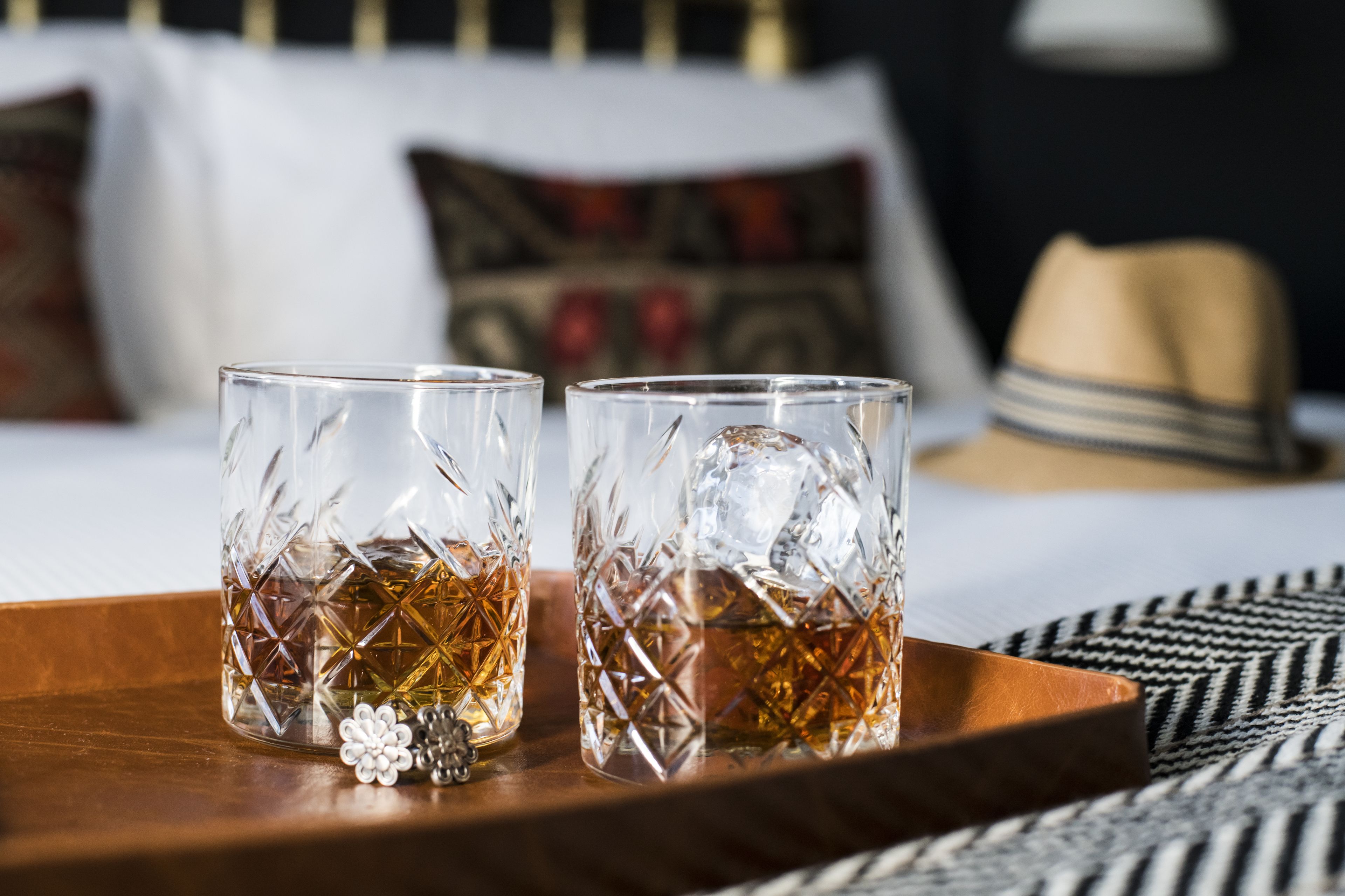Deux verres en cristal contenant des glaçons, posés sur un plateau en bois, sur un lit recouvert d’une couverture à rayures, avec un chapeau en arrière-plan.