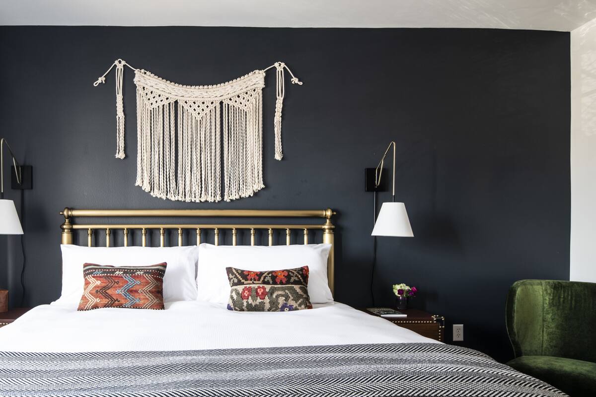 Una cama bien hecha con almohadas estampadas, un colgador de macramé en la pared y dos lámparas de noche.