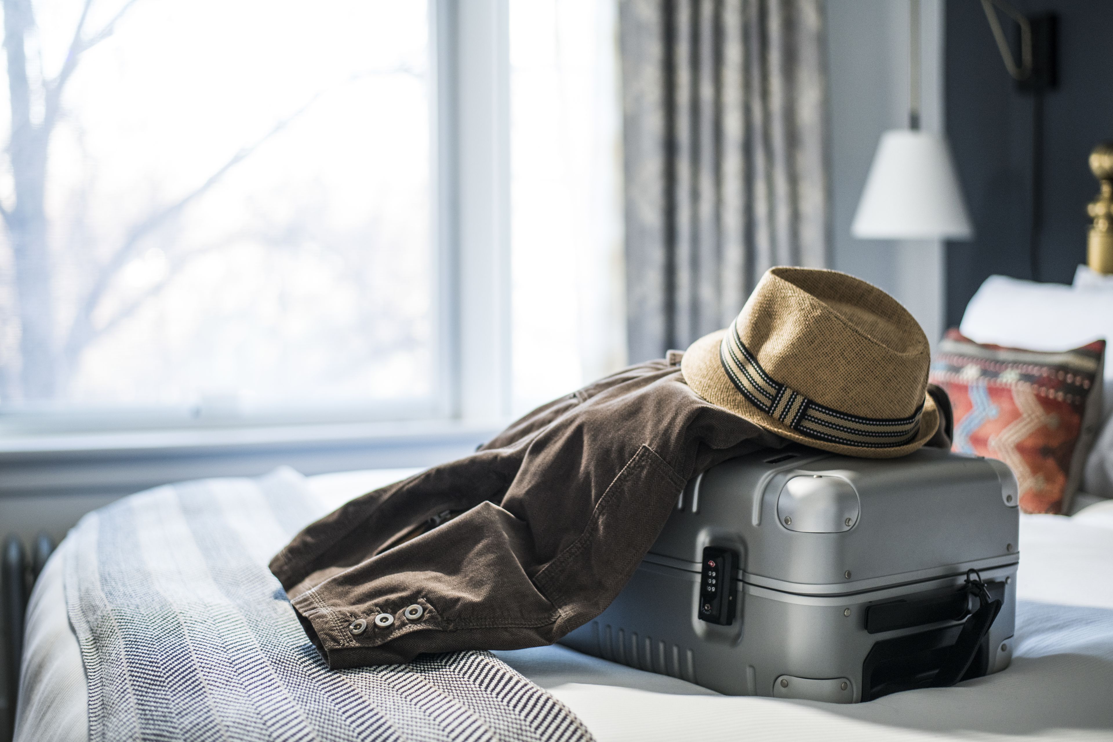 A bed with a suitcase and a hat on it, a window, and a lamp.