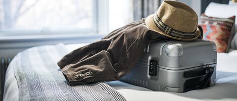 A bed with a suitcase and a hat on it, a window, and a lamp.