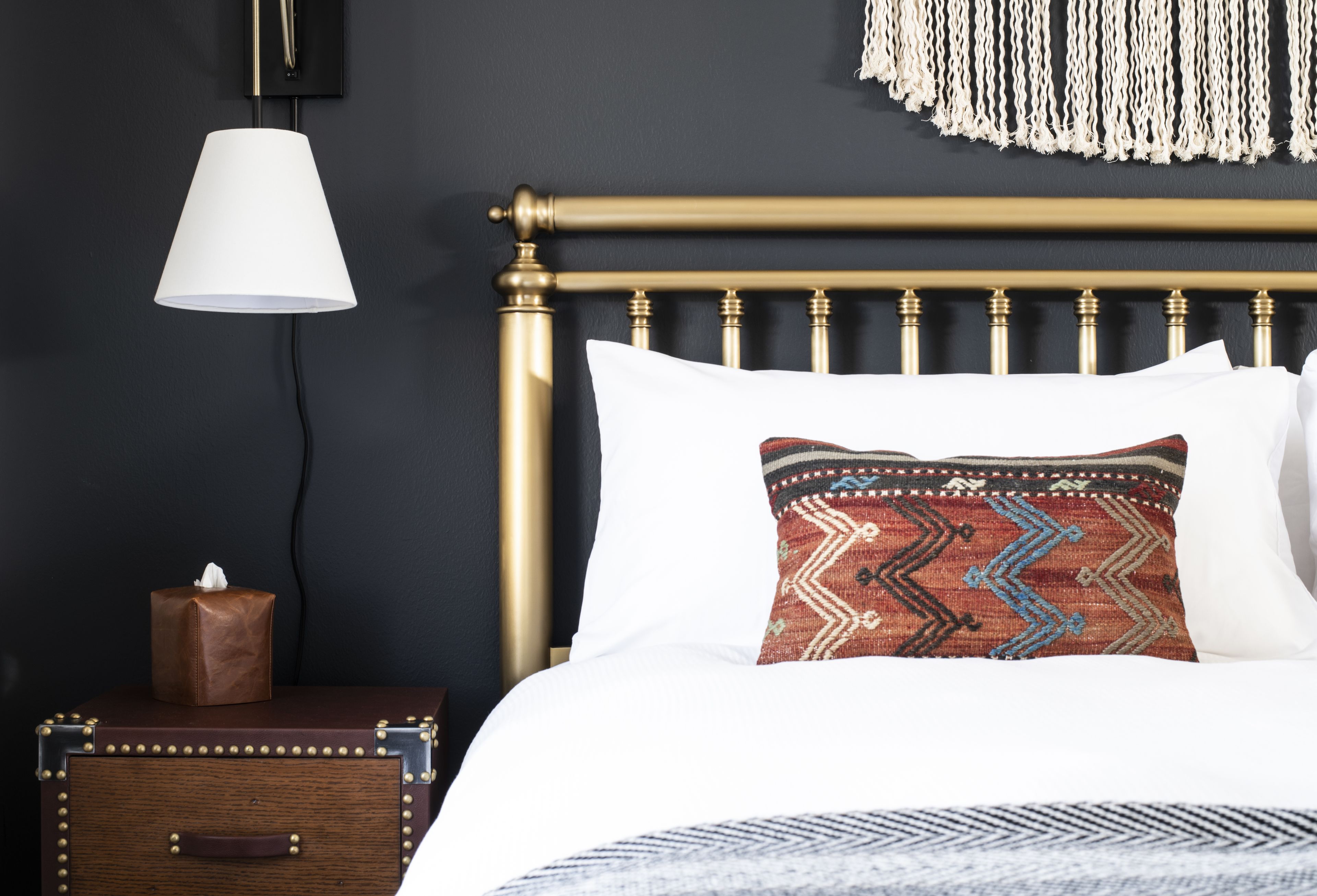 A neatly made bed with a patterned pillow, a bedside lamp, a small wooden chest, and a macrame wall hanging.