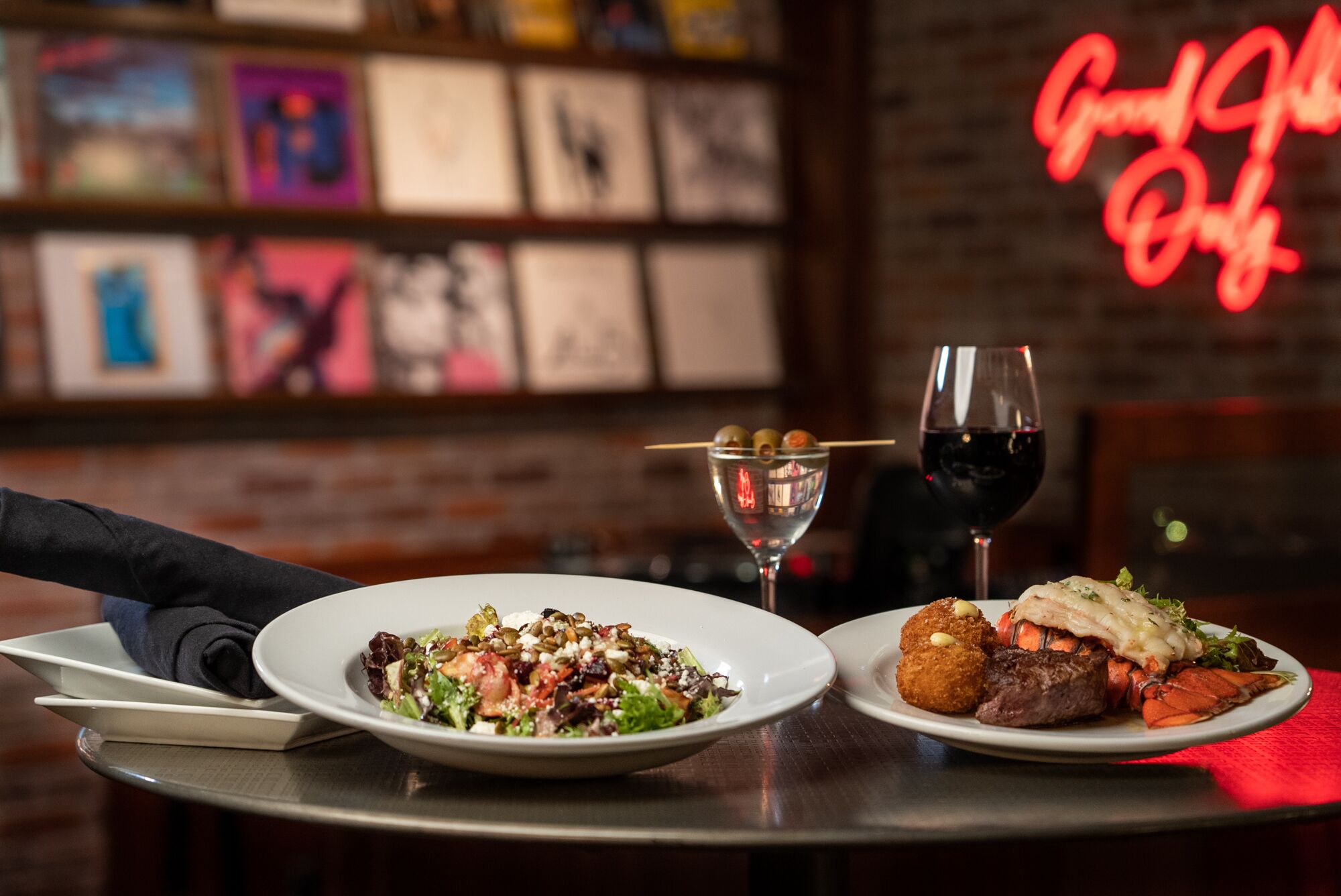 Two plates of food and two glasses of red wine on a table in a restaurant with a neon sign in the background.
