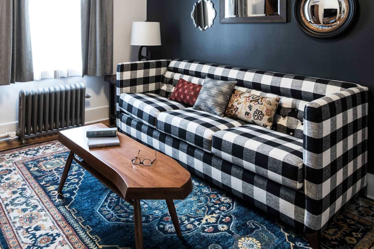 Una sala de estar con un sofá a cuadros, una mesa de centro de madera, un tapete estampado, un radiador y una ventana con cortinas.