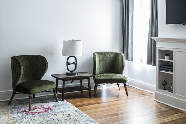 A room with two green armchairs, a small table with a lamp, a patterned rug, and a white fireplace with shelves.
