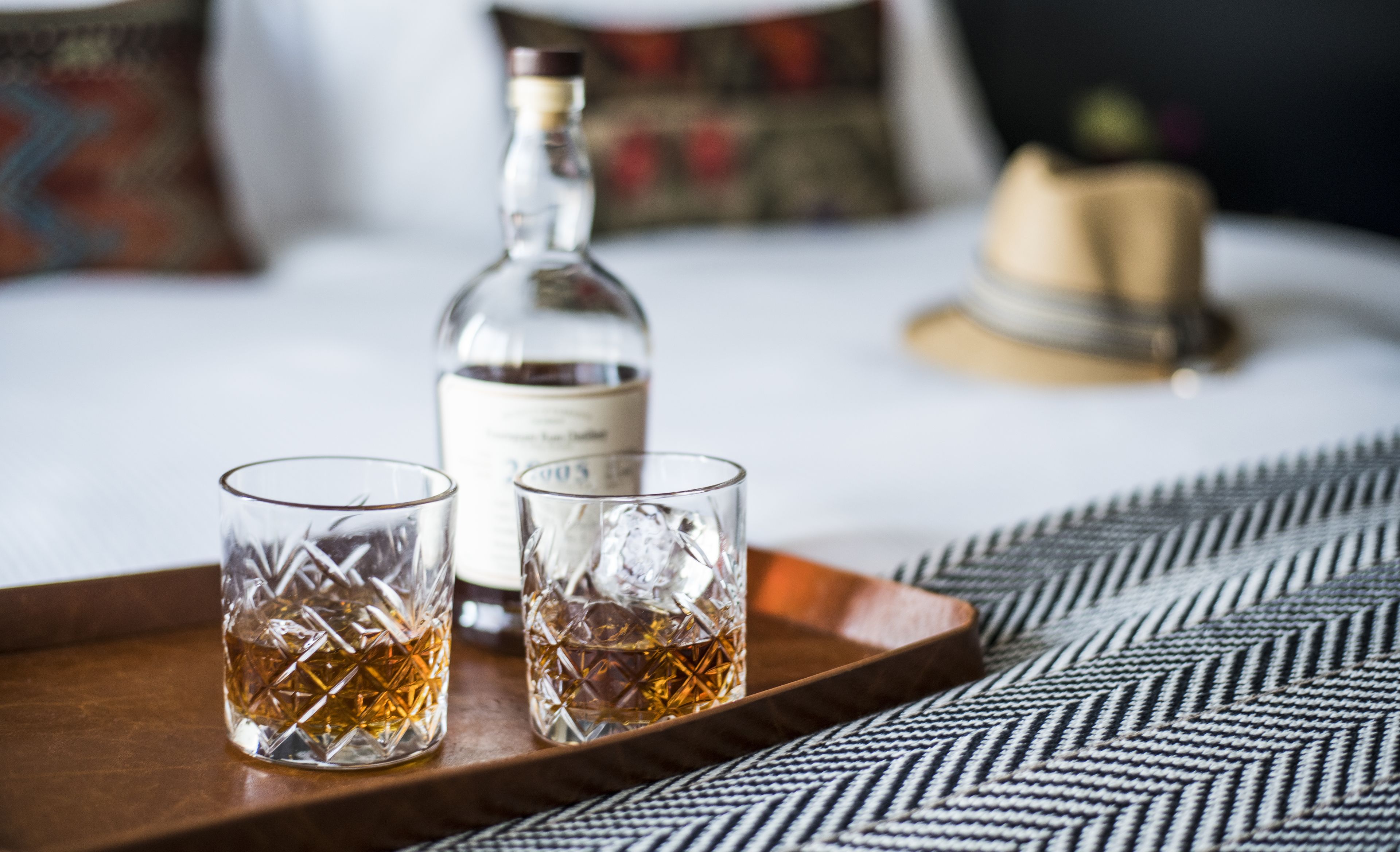 A tray with two glasses of whiskey and a bottle on a bed with a patterned bedspread.