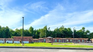 A row of single-story brick buildings with attached garages, likely a motel or small hotel, with a parking lot in front and a grassy area with a gazebo.