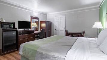 A hotel room with a bed, a desk with a computer, a chair, a microwave, a refrigerator, and a door.