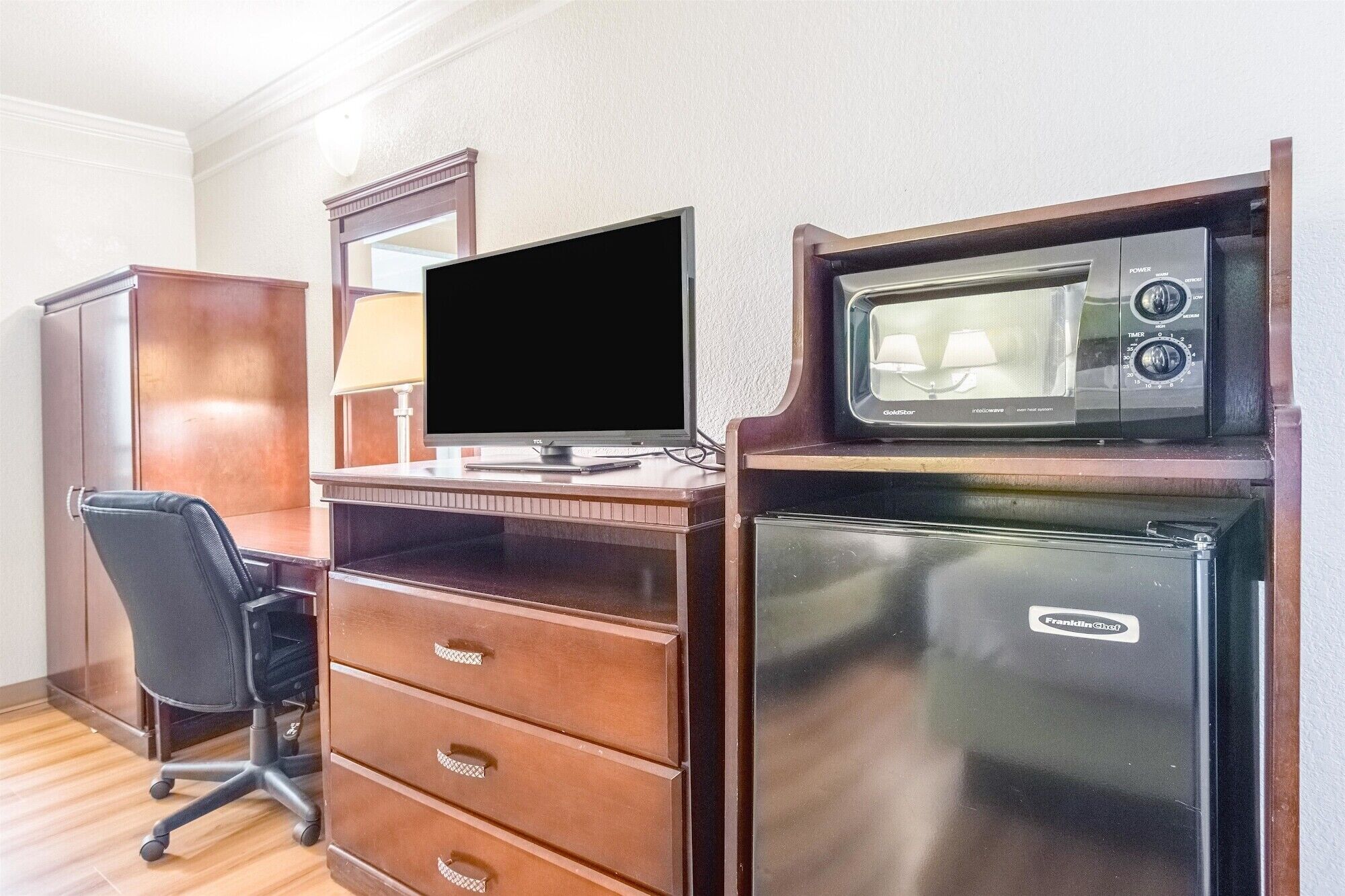 A compact hotel room with a wooden desk, a computer, a microwave, and a refrigerator.