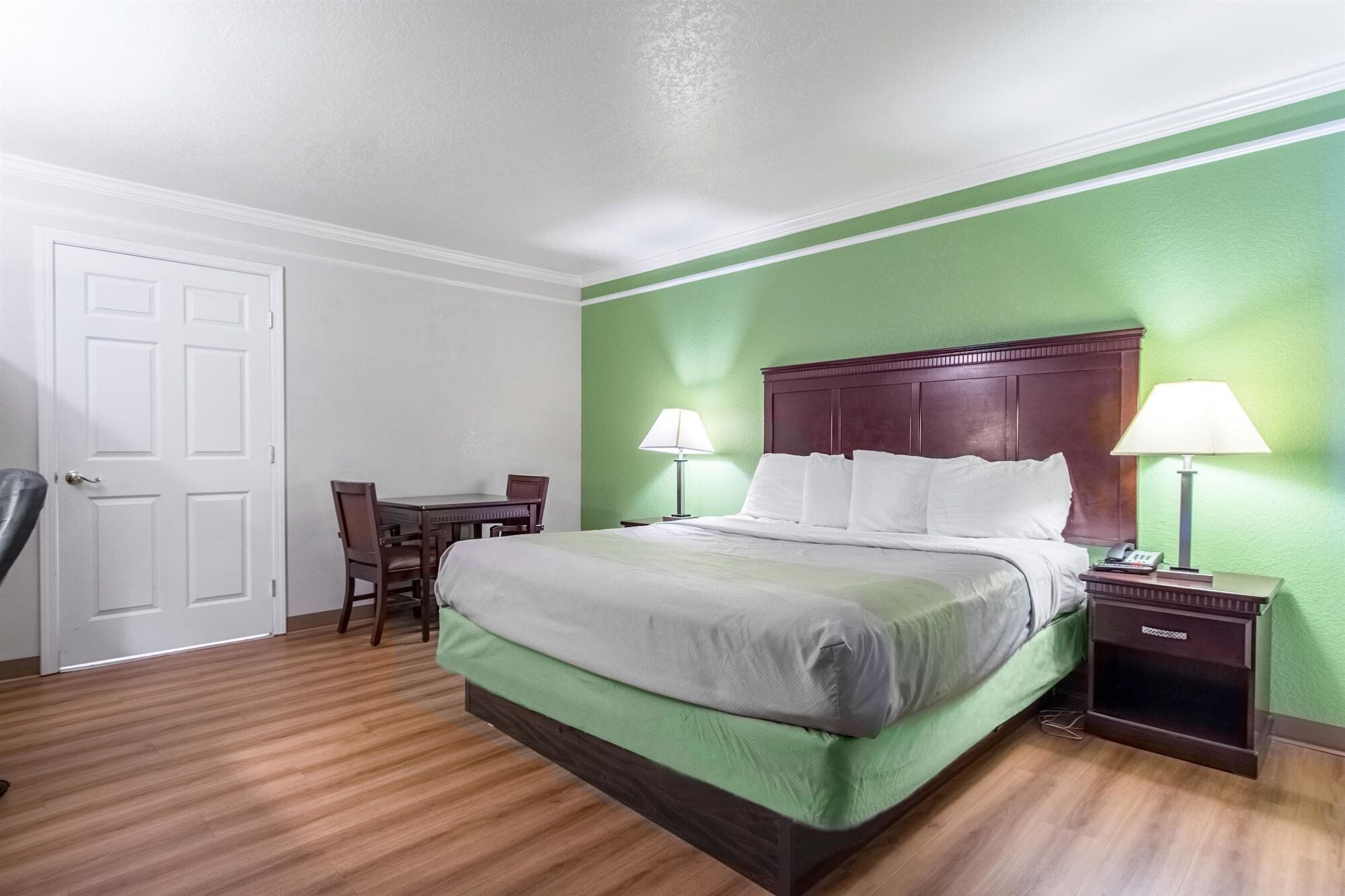 A hotel room with a green wall, a wooden floor, a bed with white bedding, a wooden headboard, two bedside tables with lamps, and a desk with a chair.