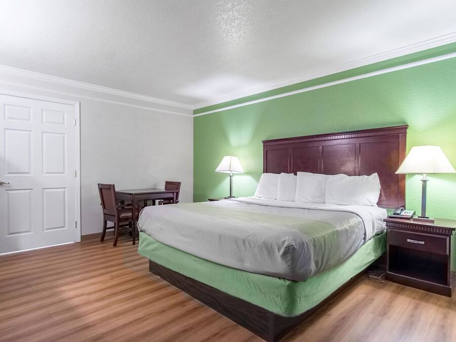 A hotel room with a green wall, a wooden floor, a bed with white bedding, a wooden headboard, two bedside tables with lamps, and a desk with a chair.