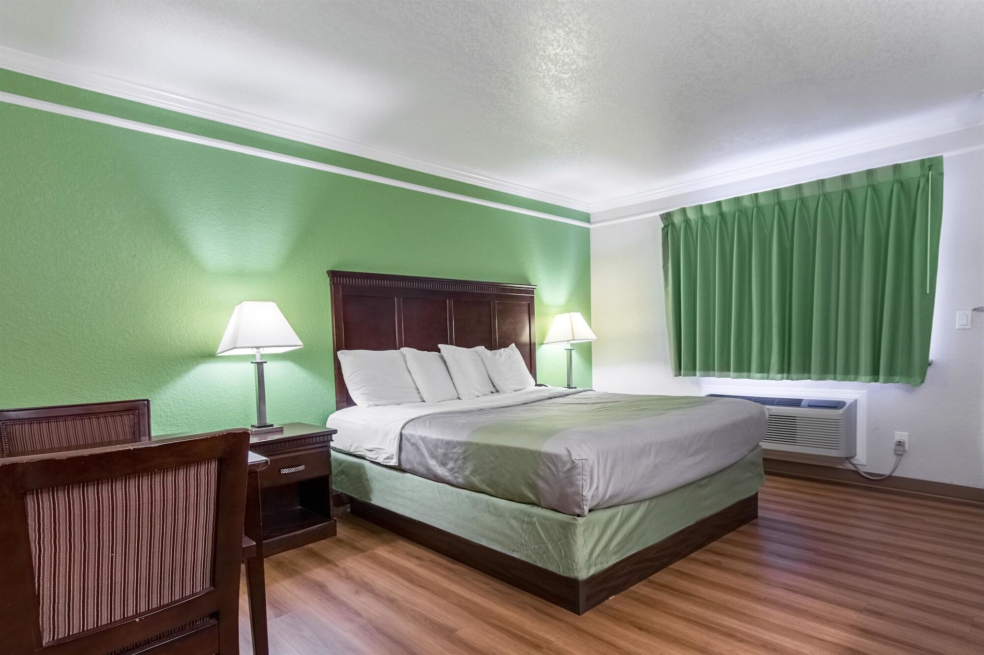 A hotel room with a bed, two bedside lamps, a nightstand, a chair, a window with green curtains, and an air conditioning unit.