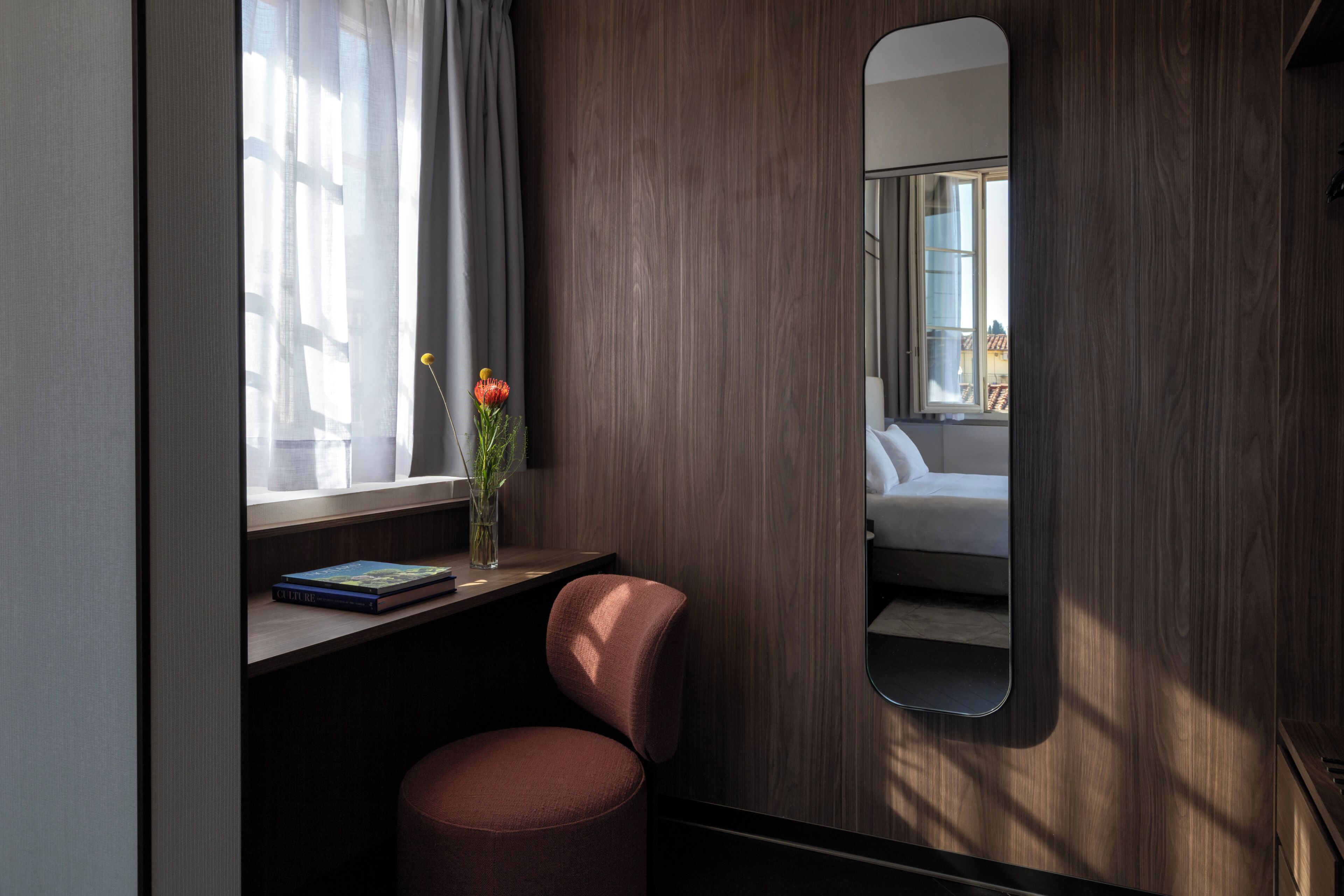 Habitación moderna con pared de madera, un espejo grande, una ventana con cortinas, un escritorio con un libro, una silla y un taburete pequeño.
