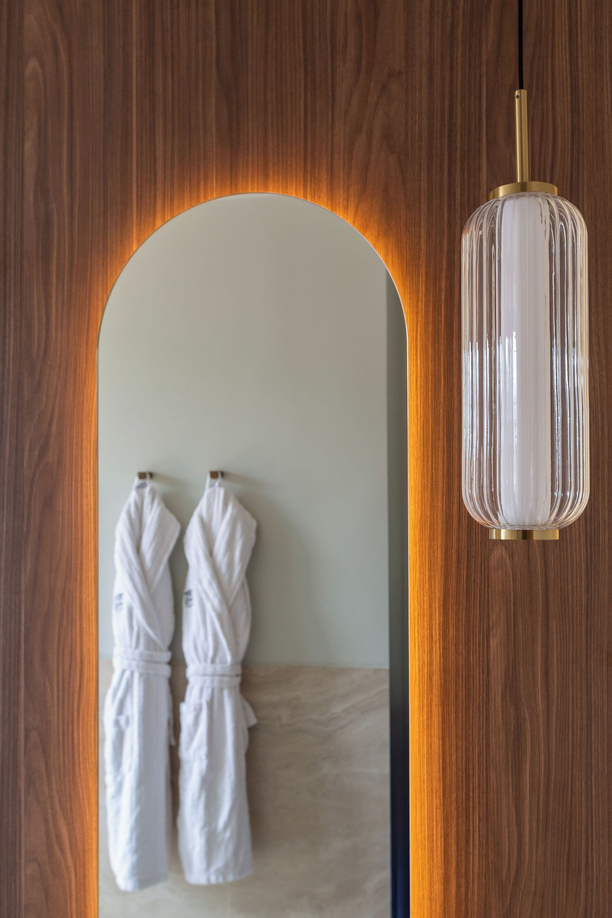 Pared de madera con un espejo que refleja dos batas blancas colgadas en un perchero.