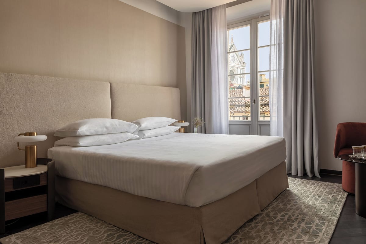 Une chambre d’hôtel avec un grand lit, une table de chevet avec une lampe, une chaise et une vue sur un immeuble par la fenêtre.