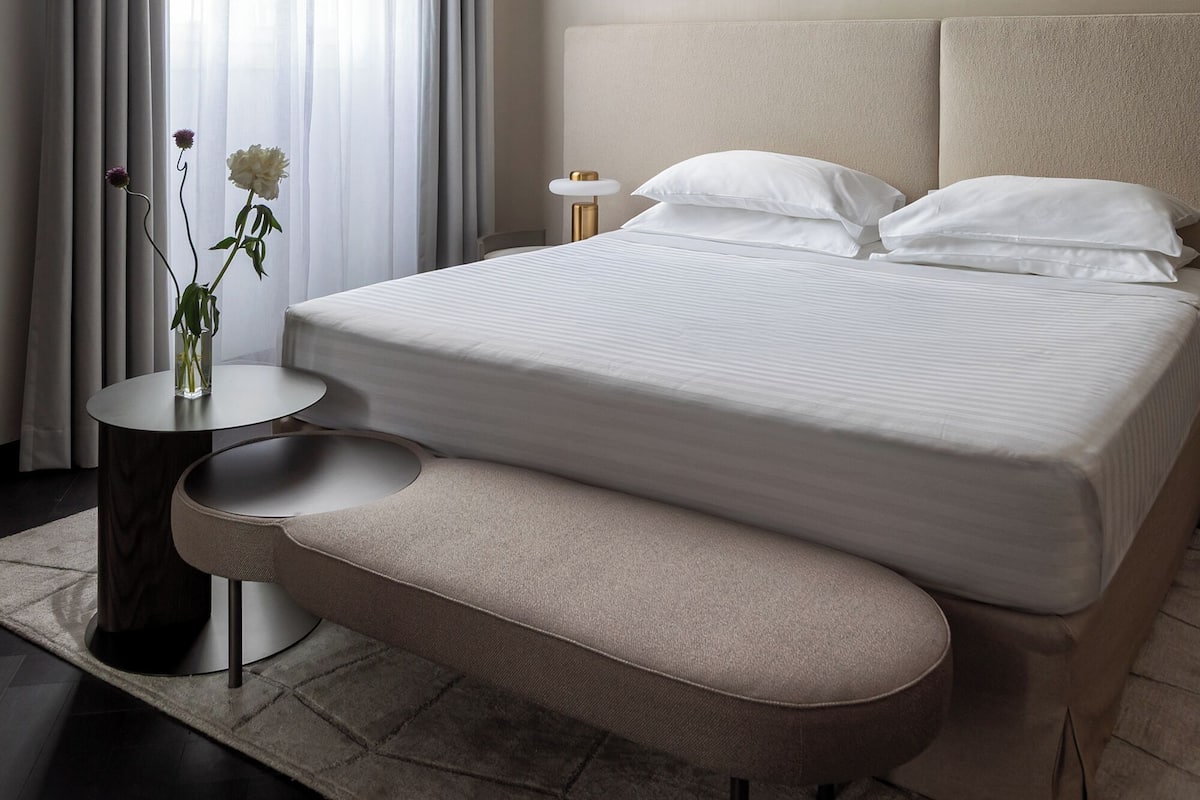 Une chambre d’hôtel moderne avec un grand lit, une table de chevet, un banc et une fenêtre avec des rideaux.