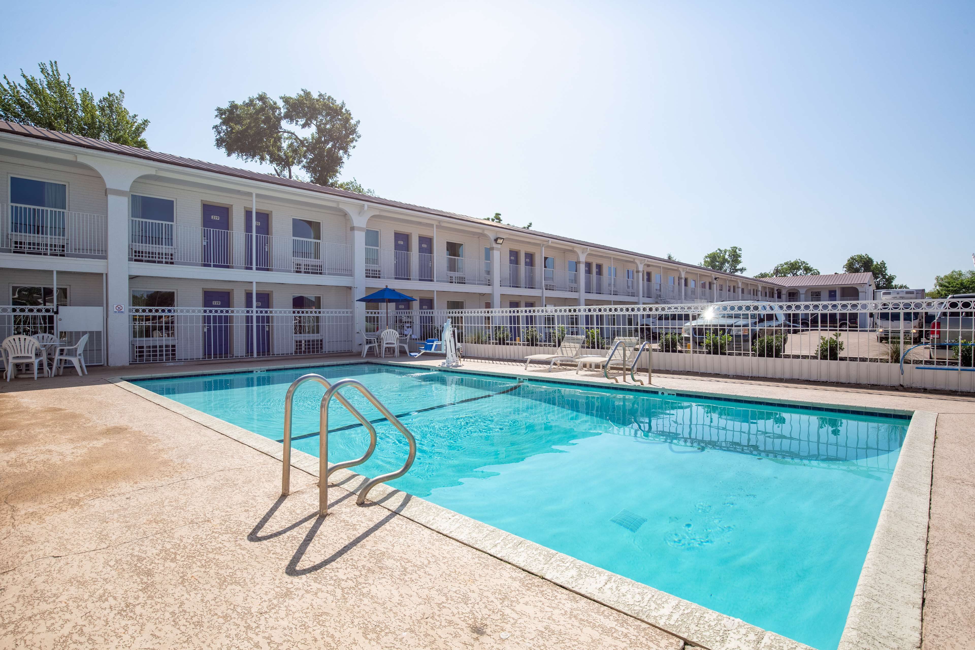 Un espace piscine avec une eau d’un bleu limpide, des chaises blanches disposées au bord de la piscine et un bâtiment doté de balcons.