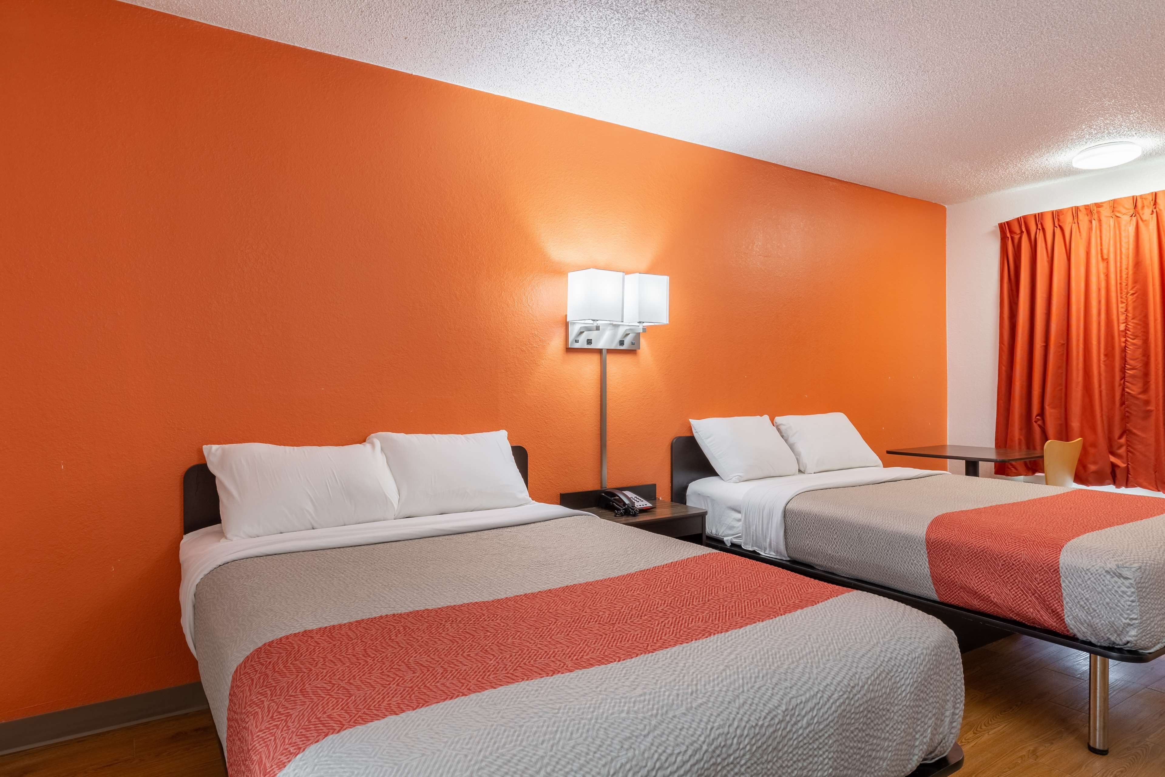 A hotel room with two beds, a nightstand, a lamp, and an orange curtain.