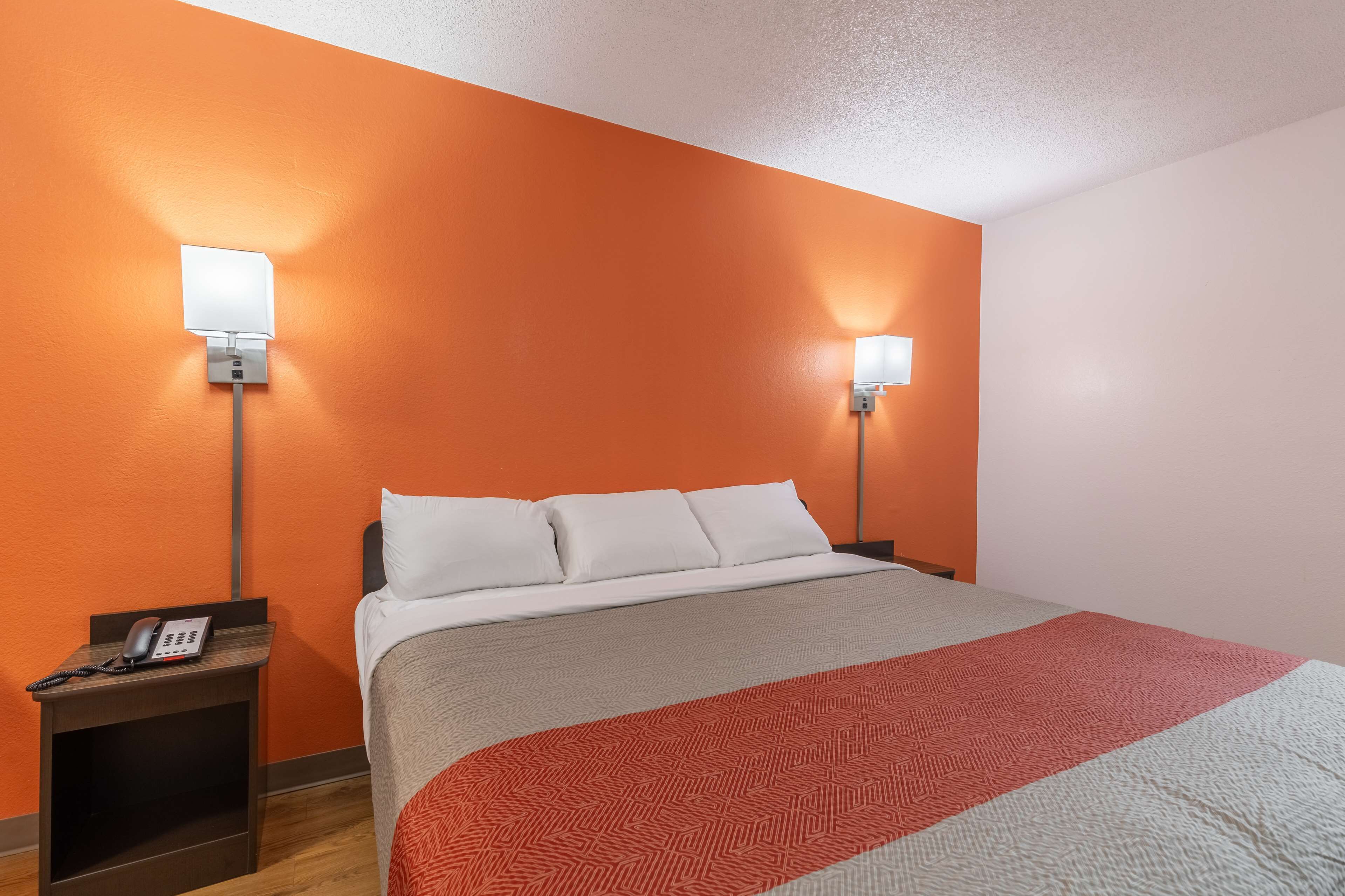 Habitación de hotel con una cama grande, dos lámparas de noche, un teléfono en una mesita y una pared naranja.